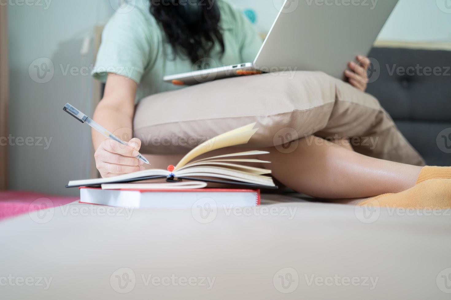 bicchieri bellissimo asiatico donna è Lavorando a partire dal casa con sua il computer portatile e Scrivi un' Nota in taccuino accanto nel il Camera da letto con un' pastello verde - Marrone colore tema. foto