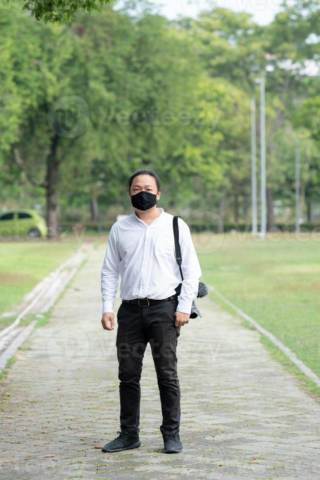 asiatico libero professionista lungo capelli uomo con nero viso maschera è in piedi e spedizione su il camminare modo nel il giardino all'aperto campo con il suo telecamera accanto lui. foto