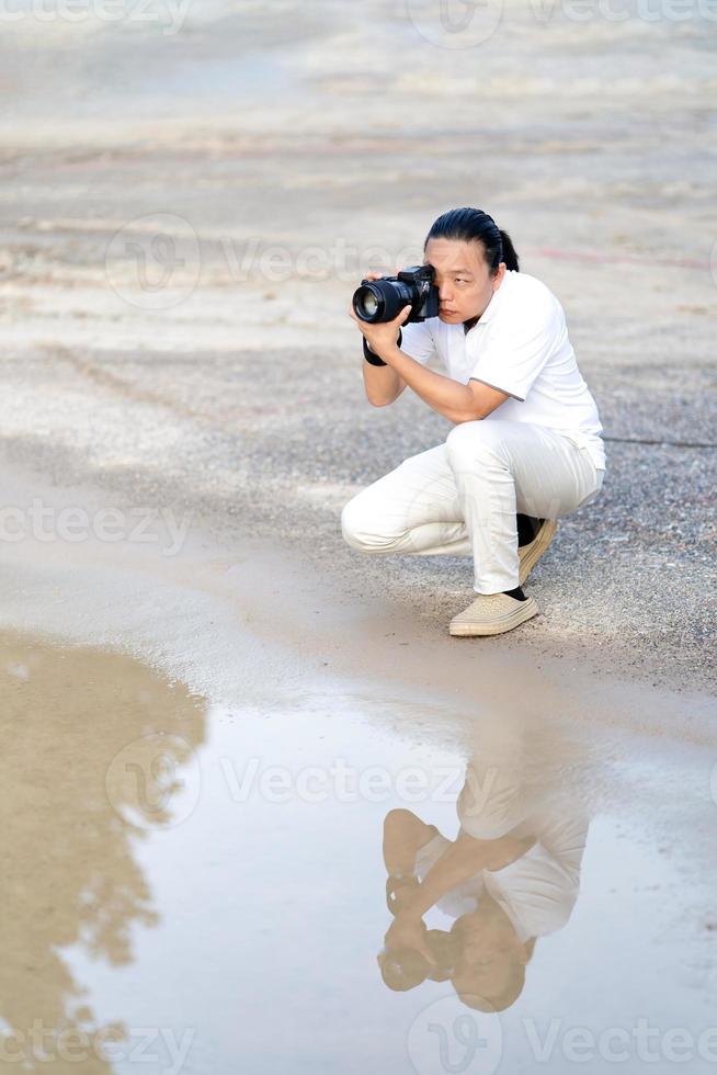 asiatico uomo detiene e grave sembra dentro il mirino mirrorless telecamera medio formato genere su cemento asfalto terra nel davanti di acqua riflesso. foto