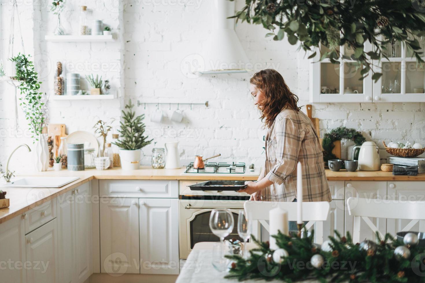 attraente sorridente donna con Riccio capelli nel plaid camicia cuoce biscotti a luminosa cucina a il casa foto