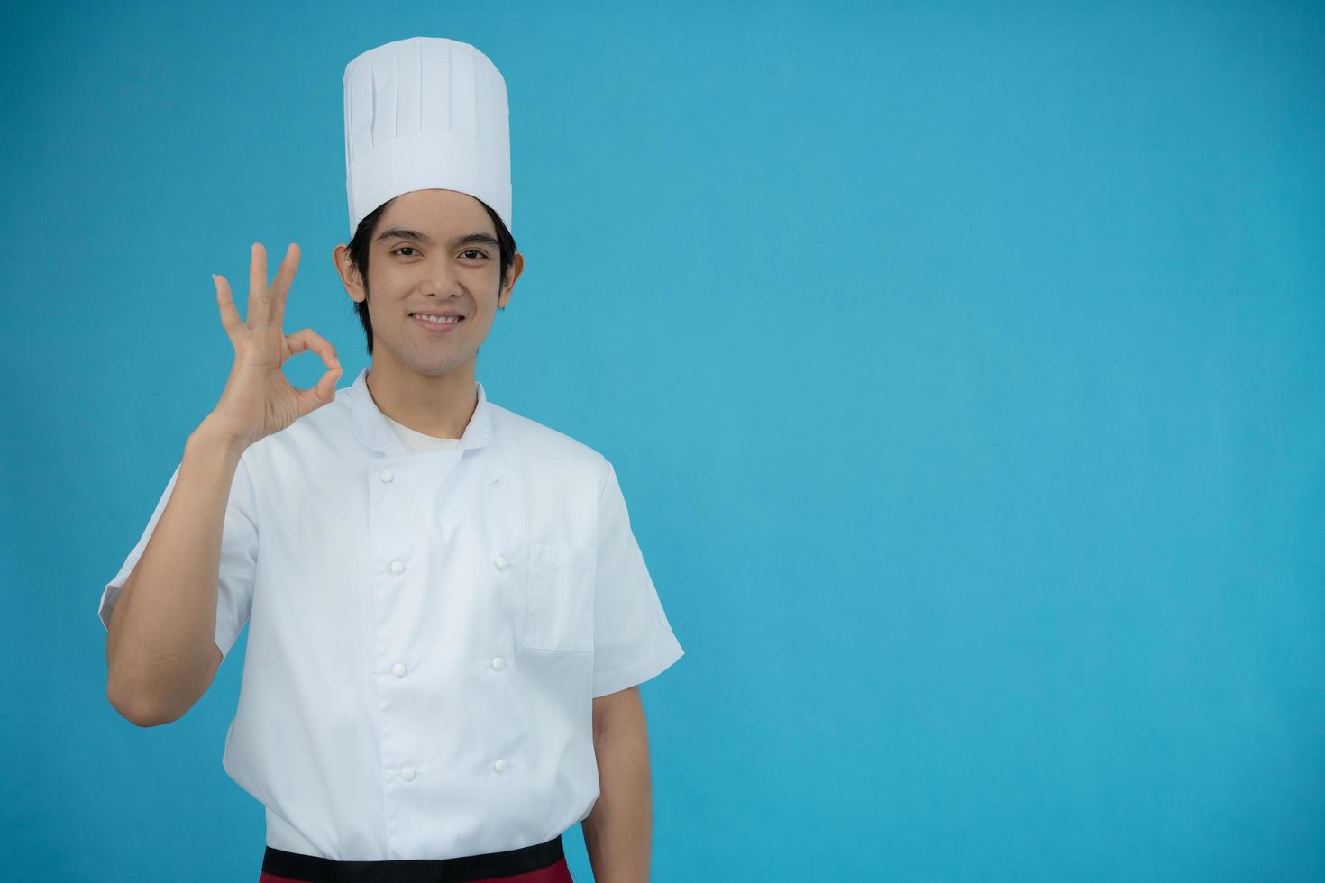 ritratto di un' bello asiatico uomo indossare capocuoco Abiti su blu isolato sfondo. capocuoco sorrisi e è contento con il cibo quello è ok con il presentazione. foto