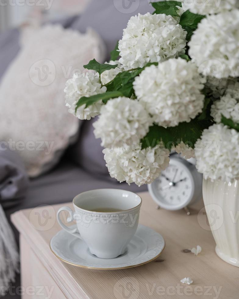 un' tazza di caffè, un allarme orologio e un' vaso di bianca fiori su il capezzale tavolo. tè volta, caffè tempo foto