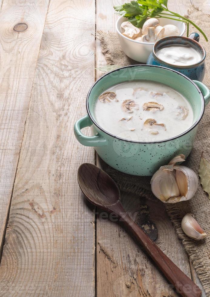 turchese ceramica padella con fungo crema la minestra, tazza con acida crema, ciotola con fresco champignons, cucchiaio, aglio su tela ruvida su di legno sfondo. foto