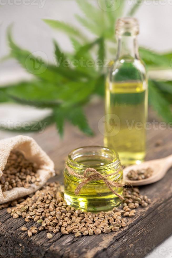 canapa olio nel Due bicchiere vaso e bottiglia con grano nel il sacco su di legno tavola con le foglie di canapa su sfondo. foto