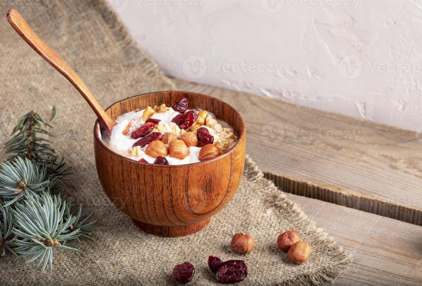 di legno ciotola di fatti in casa Yogurt con lanciato avena noccioline, secco mirtilli tela ruvida su di legno tavole vicino bianca parete. foto