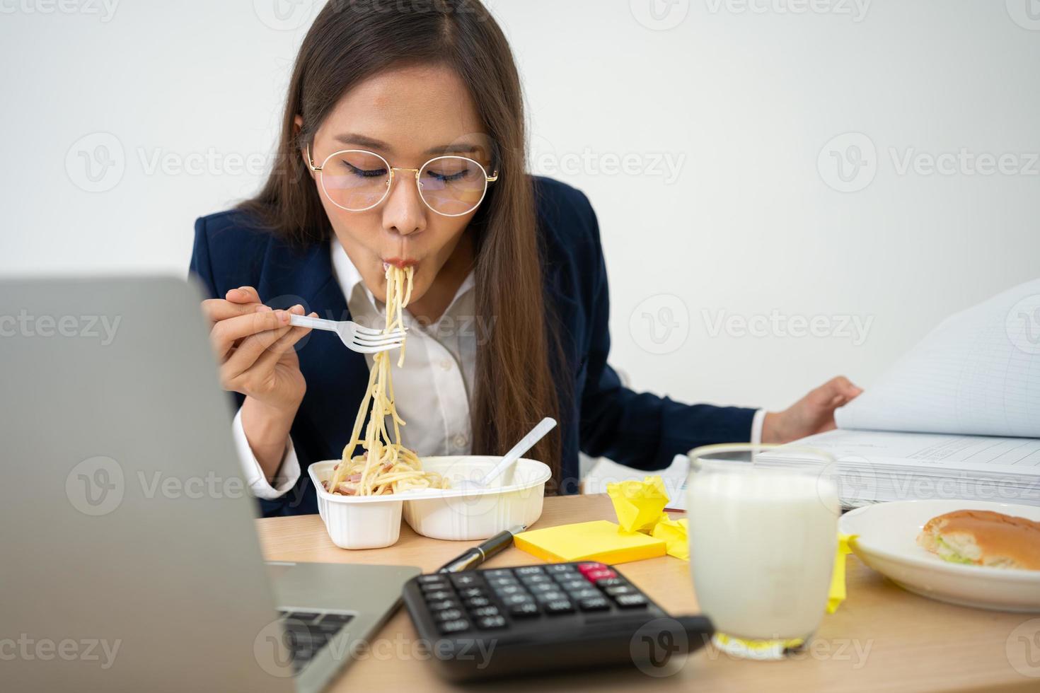 occupato e stanco donna d'affari mangiare spaghetti per pranzo a il scrivania ufficio e Lavorando per consegnare finanziario dichiarazioni per un' capo. oberati di lavoro e malsano per pronto pasti, bruciato concetto. foto