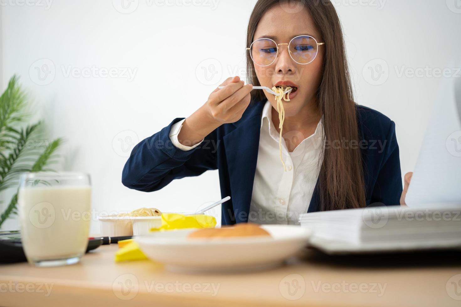 occupato e stanco donna d'affari mangiare spaghetti per pranzo a il scrivania ufficio e Lavorando per consegnare finanziario dichiarazioni per un' capo. oberati di lavoro e malsano per pronto pasti, bruciato concetto. foto