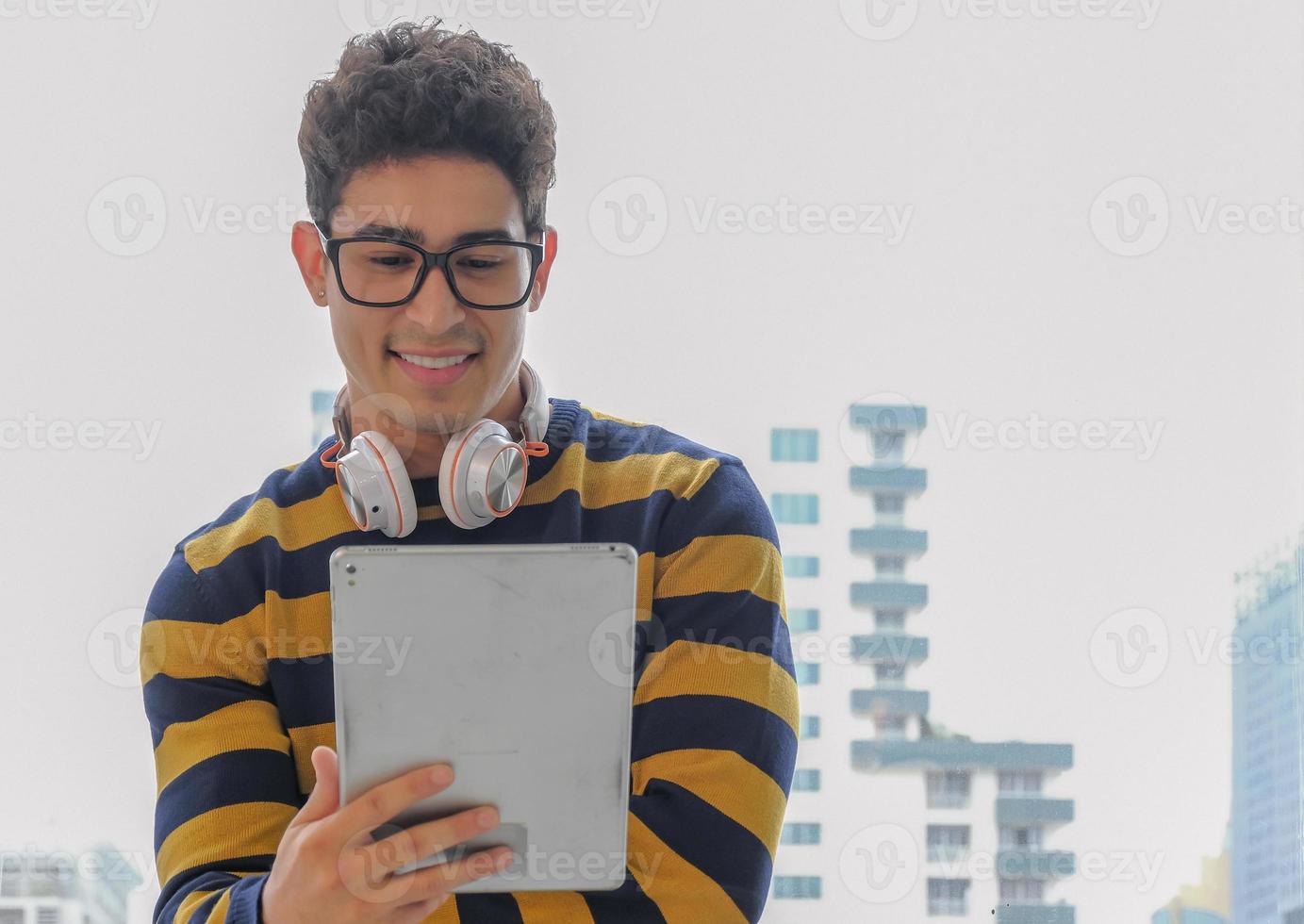 giovane creativo attività commerciale bicchieri uomo indossare cuffie e sensazione contento mentre guardare a tavoletta dispositivo per Lavorando con sorridente viso nel davanti di ufficio finestra sfondo. indietro leggero fotografia. foto