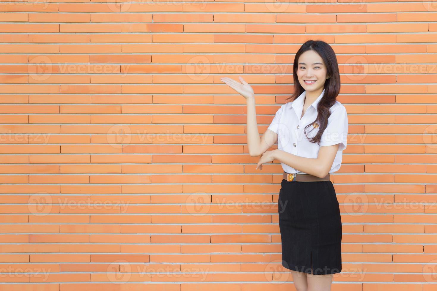 ritratto di un adulto tailandese alunno nel Università alunno uniforme. asiatico bellissimo ragazza in piedi per presente qualcosa con fiducia su mattone muri sfondo. foto