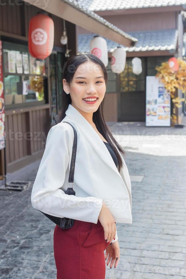 giovane asiatico attività commerciale Lavorando donna indossare bianca completo da uomo e rosso pantaloni sorridente, portare Borsa mentre a piedi nel il città. foto