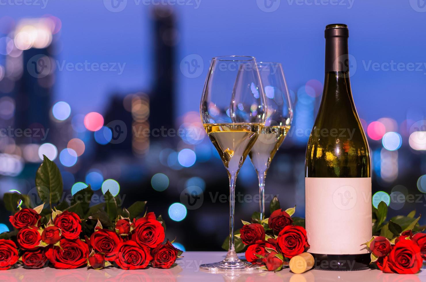 selettivo su primo bicchiere di bianca vino con sfocato bottiglia e rosso Rose su colorato bokeh leggero a partire dal città sfondo per san valentino cenare concetto. foto