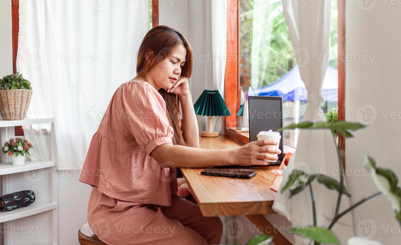 un' contento femmina a un' bar utilizzando un' il computer portatile nel mano e un' carta tazza di caffè. giovane bianca donna con lungo capelli seduta nel un' caffè negozio occupato Lavorando su sua il computer portatile. foto