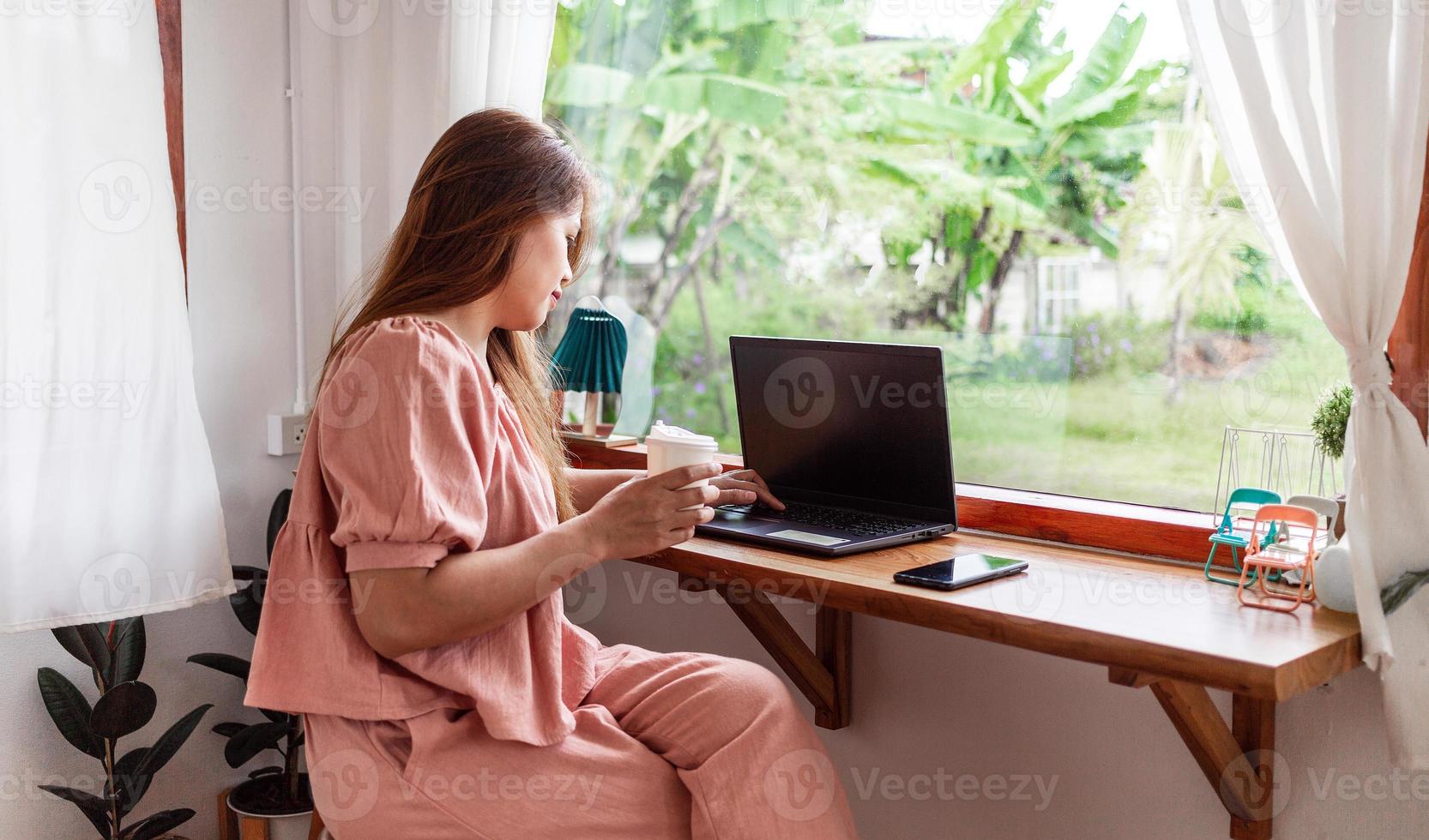 un' contento femmina a un' bar utilizzando un' il computer portatile nel mano e un' carta tazza di caffè. giovane bianca donna con lungo capelli seduta nel un' caffè negozio occupato Lavorando su sua il computer portatile. foto