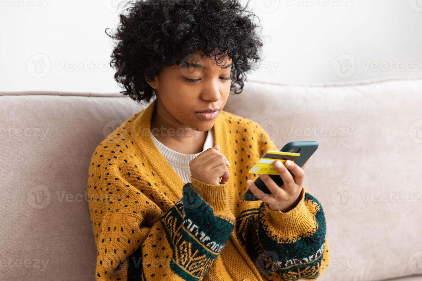 africano americano donna shopping in linea Tenere smartphone pagare con oro credito carta. ragazza seduta a casa acquisto su Internet accedere credito carta particolari. in linea shopping e-commerce consegna servizio. foto