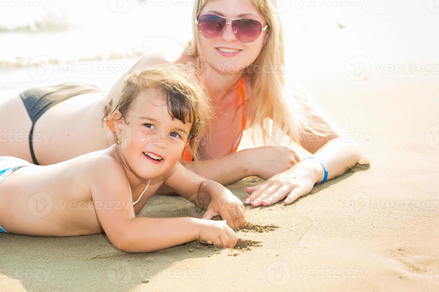 ritratto di bellissimo giovane genitori e carino poco ragazza nel costumi da bagno e sole bicchieri sorridente, dire bugie su il soleggiato spiaggia. genitori siamo guardare a loro figlia foto