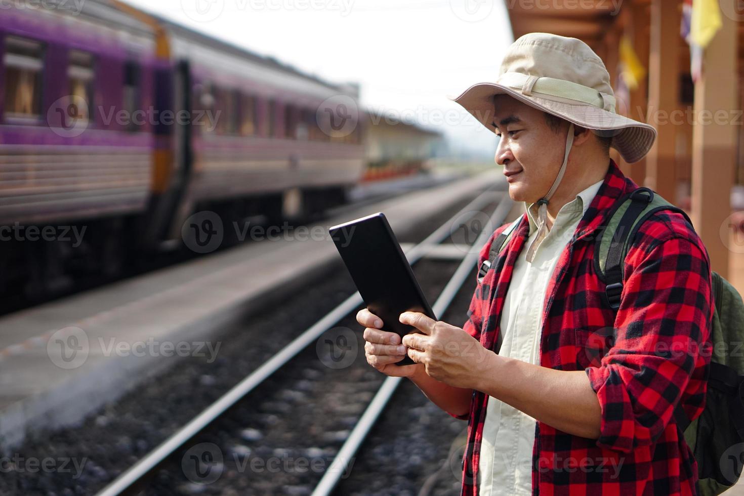 asiatico uomo viaggiatore è a ferrovia stazione, indossa cappello, detiene inteligente tavoletta. concetto, viaggio di treno nel Tailandia può libro o di biglietto in linea. tecnologia e trasporto. foto