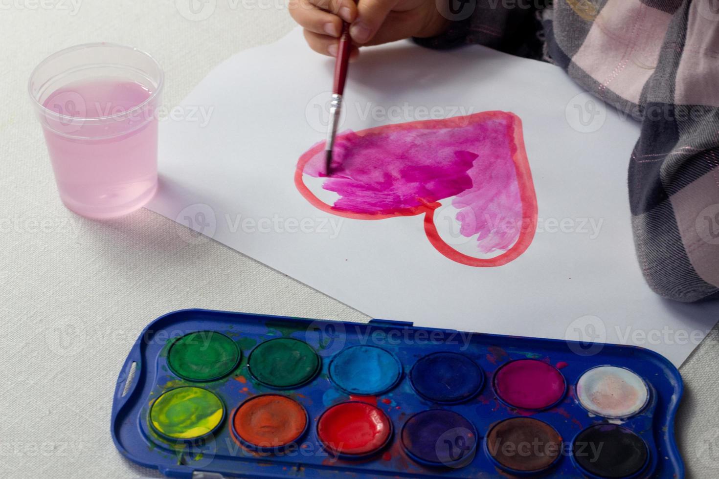 poco ragazza seduta a il tavolo e disegno rosso cuore acquerello, san valentino giorno cartolina, saluto carta, arte scuola concetto, pubblicità, bambino pittura acquerello a casa, bambino attività, bambini tempo libero. foto