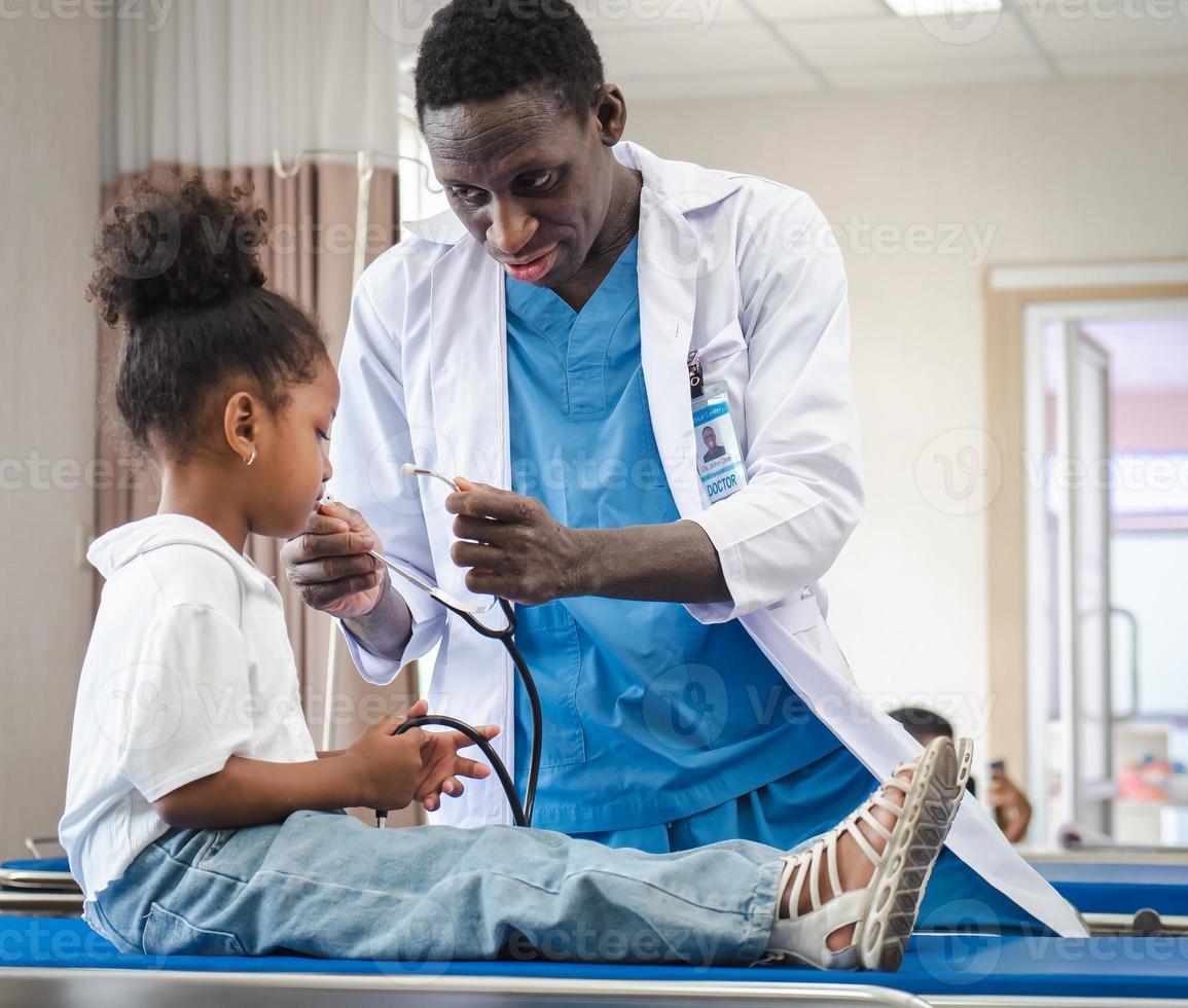 messa a fuoco selettiva al medico persona che gioca con un simpatico bambino afro paziente nel reparto ospedaliero. amichevole pediatra divertente ragazza ragazzo godendo con lo stetoscopio per il battito cardiaco durante l'esame medico. foto