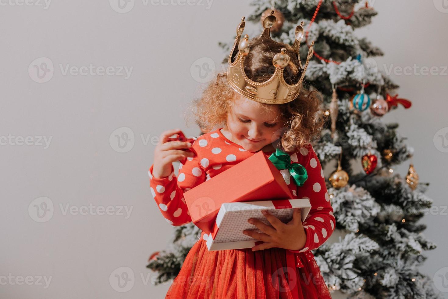 poco Riccio ragazza nel un' rosso carnevale vestito con un' Santa Stampa e un' corona è giocando con un' giocattolo cervo Il prossimo per il Natale albero. stile di vita, figli di emozioni. spazio per testo. alto qualità foto