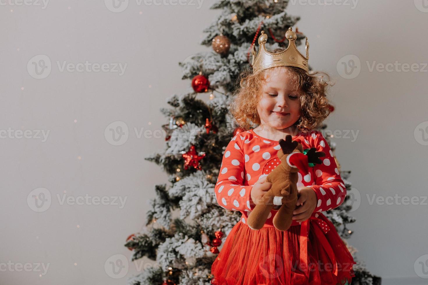 poco Riccio ragazza nel un' rosso carnevale vestito con un' Santa Stampa e un' corona è giocando con un' giocattolo cervo Il prossimo per il Natale albero. stile di vita, figli di emozioni. spazio per testo. alto qualità foto