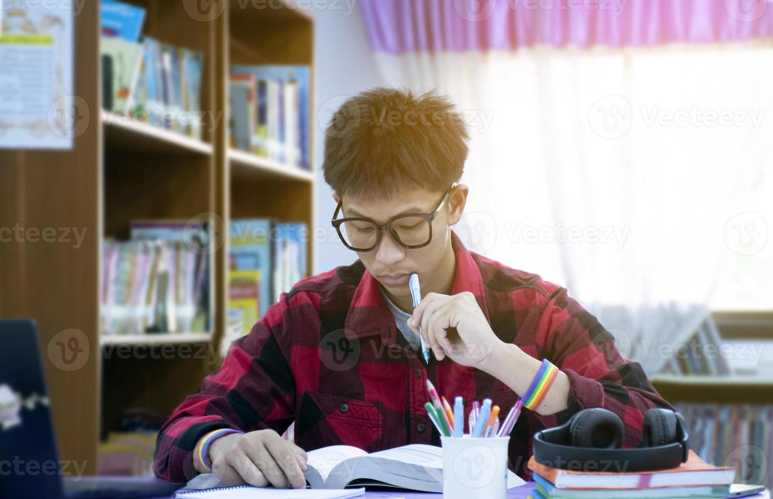 ritratto giovane asiatico ragazzo indossare occhiali e arcobaleno braccialetto, Tenere penna, seduta nel biblioteca, lettura libri e concentrandosi per libro su tavolo prima medio termine test e finale test Il prossimo giorno. foto