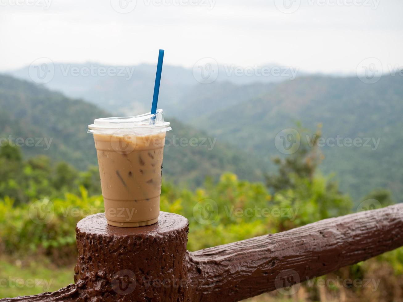 un' bicchiere di ghiacciato caffè è posto su il recinto contro un' fondale di montagne e cielo. foto