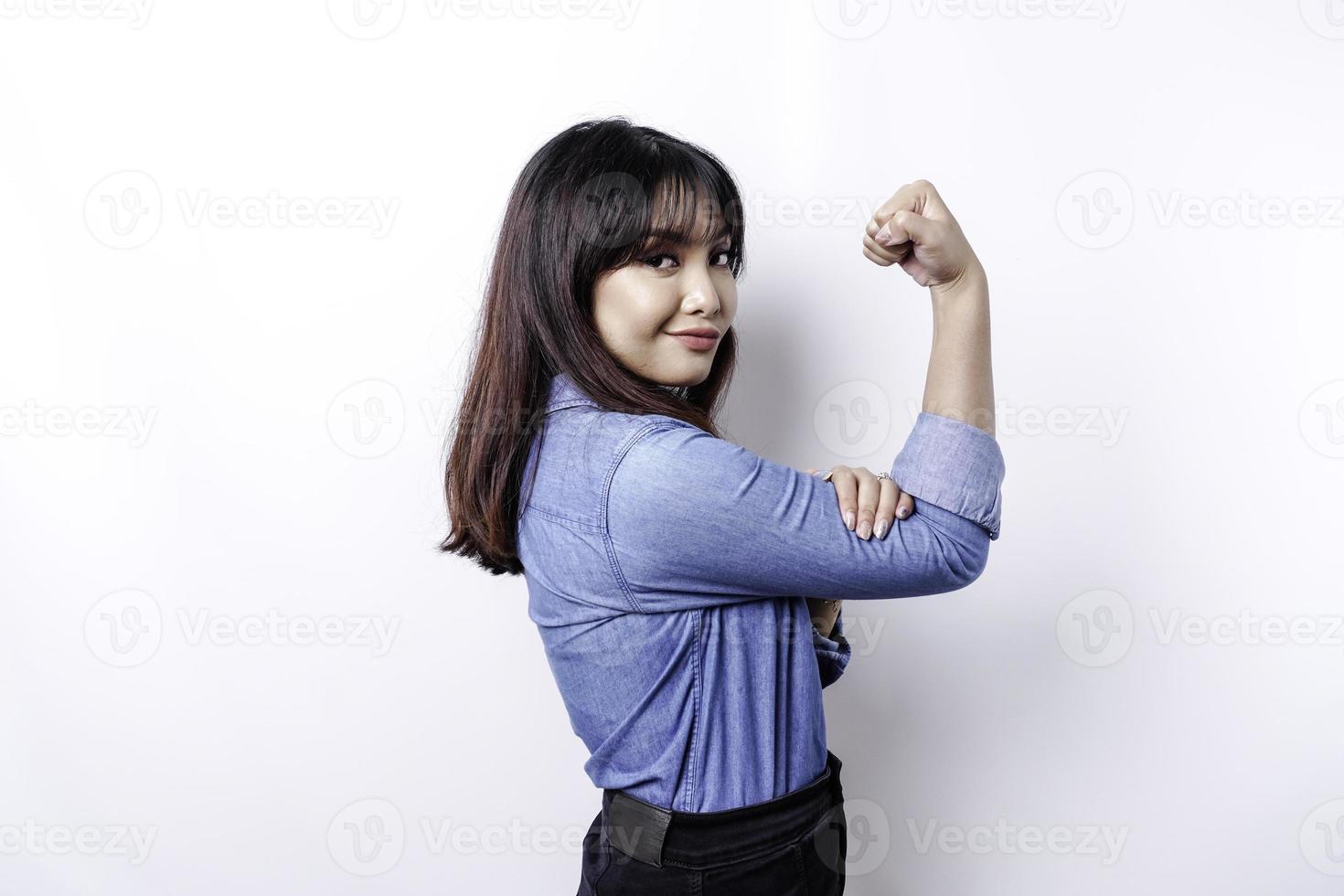 eccitato asiatico donna indossare un' blu camicia mostrando forte gesto di sollevamento sua braccia e muscoli sorridente con orgoglio foto