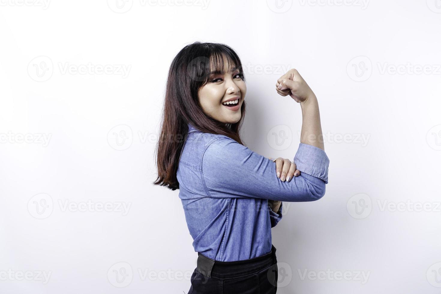 eccitato asiatico donna indossare un' blu camicia mostrando forte gesto di sollevamento sua braccia e muscoli sorridente con orgoglio foto