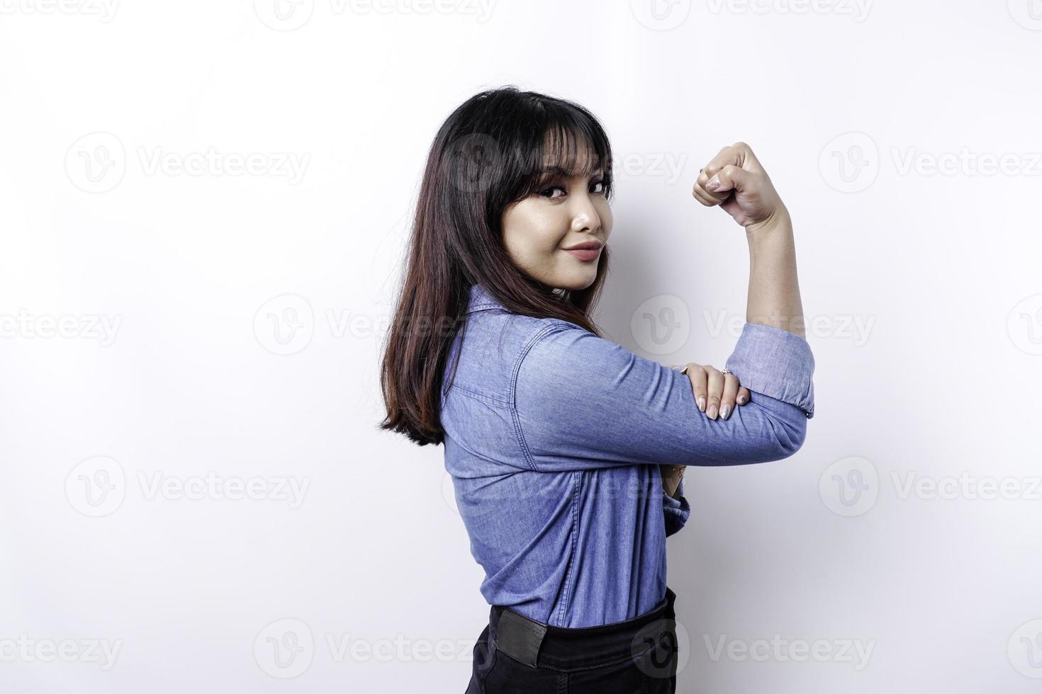 eccitato asiatico donna indossare un' blu camicia mostrando forte gesto di sollevamento sua braccia e muscoli sorridente con orgoglio foto