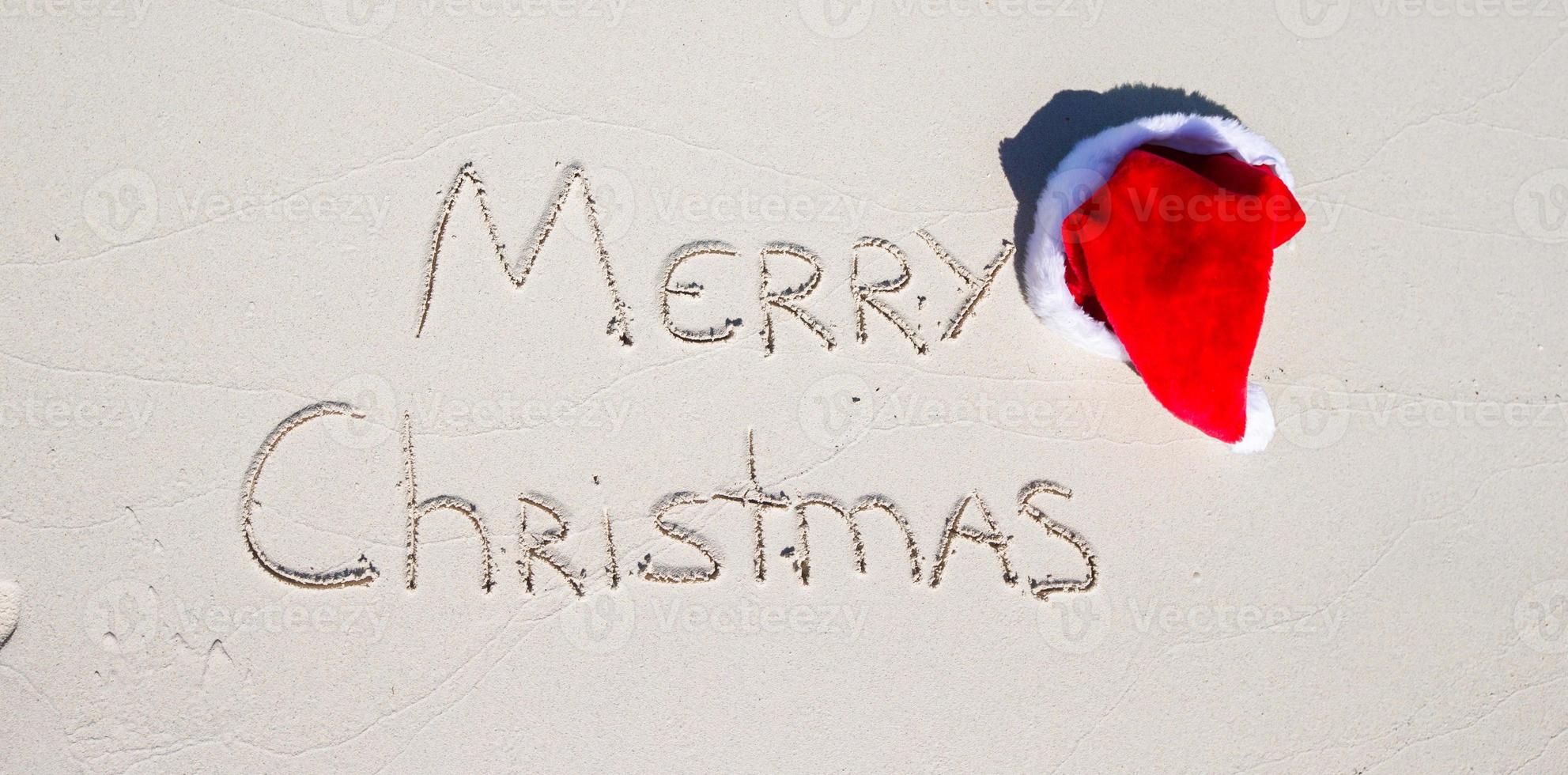 Santa cappello su bianca sabbioso spiaggia e allegro Natale scritto nel il sabbia foto