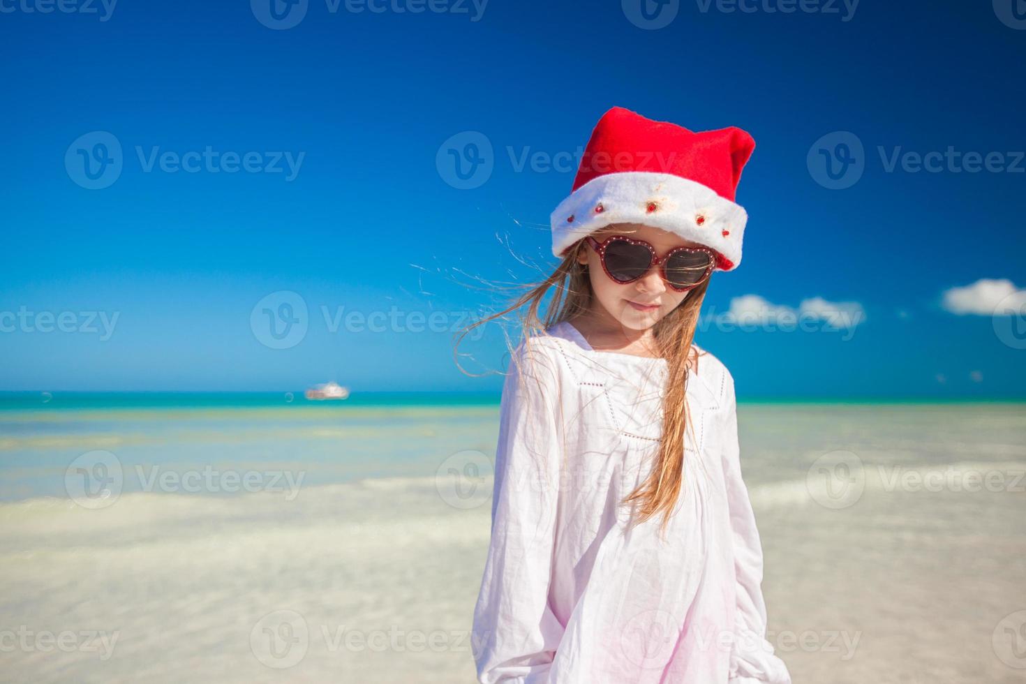 poco ragazza nel rosso cappello Santa Claus e occhiali da sole su il spiaggia foto