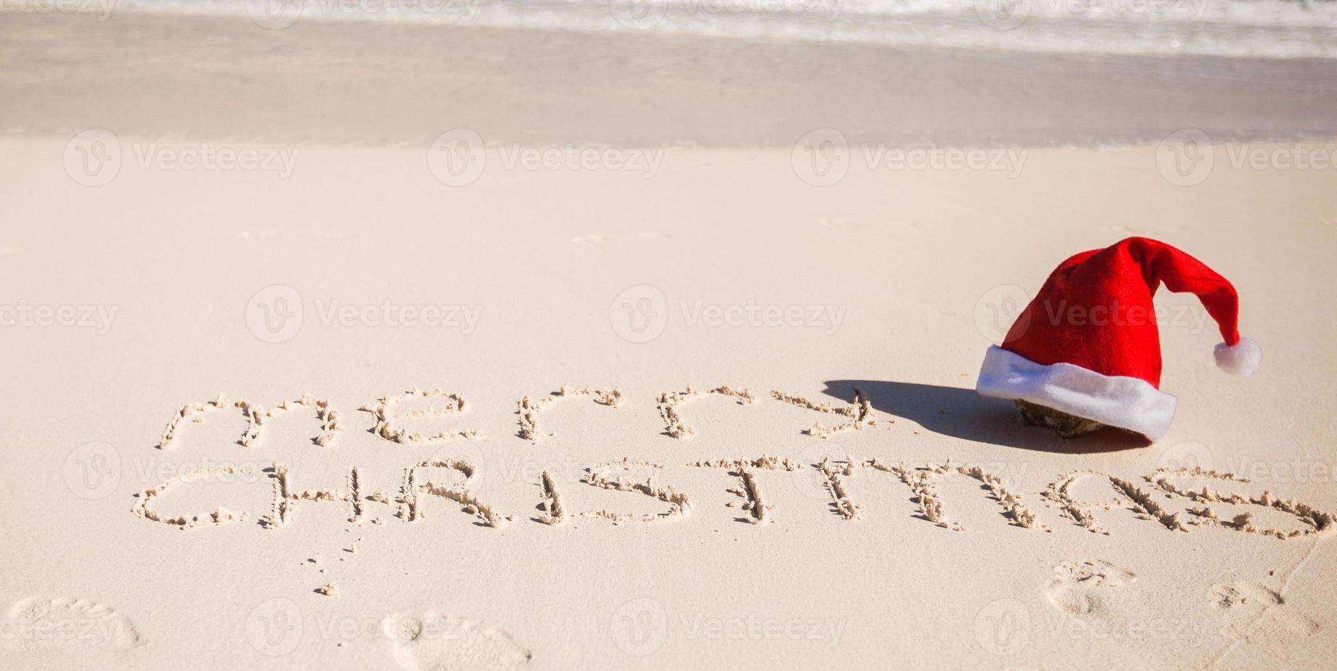 Santa cappello su bianca sabbioso spiaggia e allegro Natale scritto nel il sabbia foto