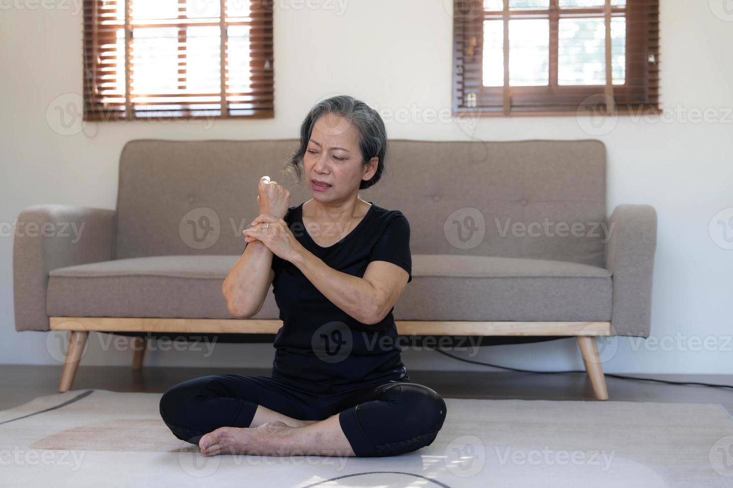 60s anziano asiatico donna massaggio sua polso, sensazione dolore e rigonfiamento nel il articolazioni, ferito mano durante yoga pratica a casa. foto