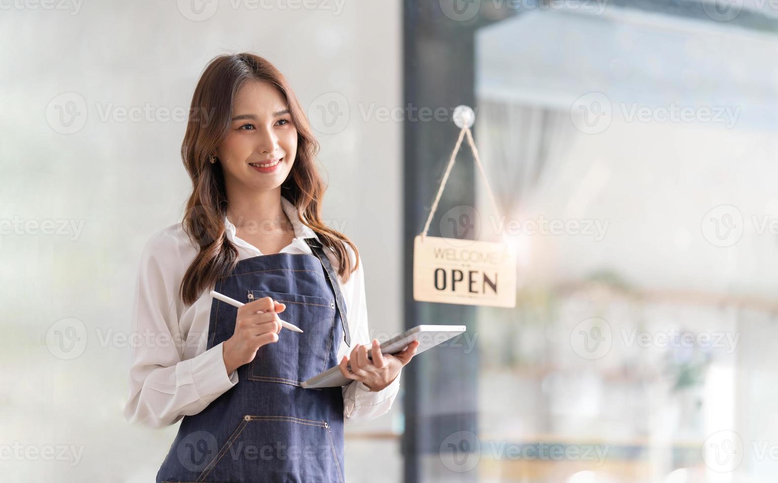 bellissimo asiatico giovane barista donna nel grembiule Tenere tavoletta e in piedi nel davanti di il porta di bar con Aperto cartello tavola. attività commerciale proprietario avviare concetto. foto