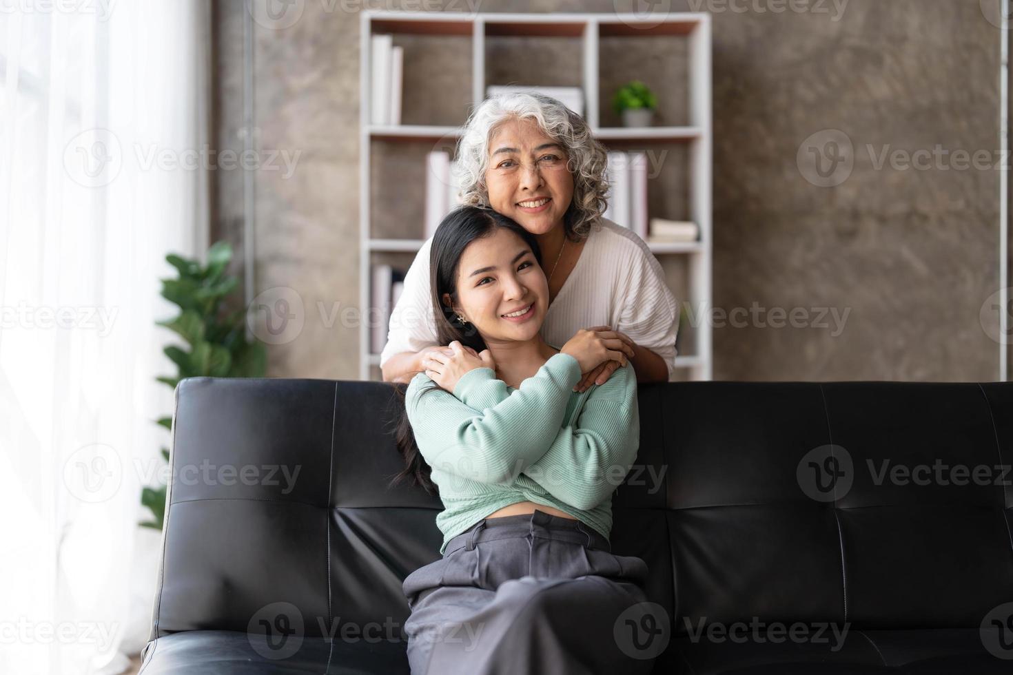 amorevole adulto figlia abbracciare più vecchio madre, in piedi dietro a divano a casa, famiglia godendo tenero momento insieme, giovane donna e maturo mamma o nonna guardare a ogni Altro, Due generazioni foto