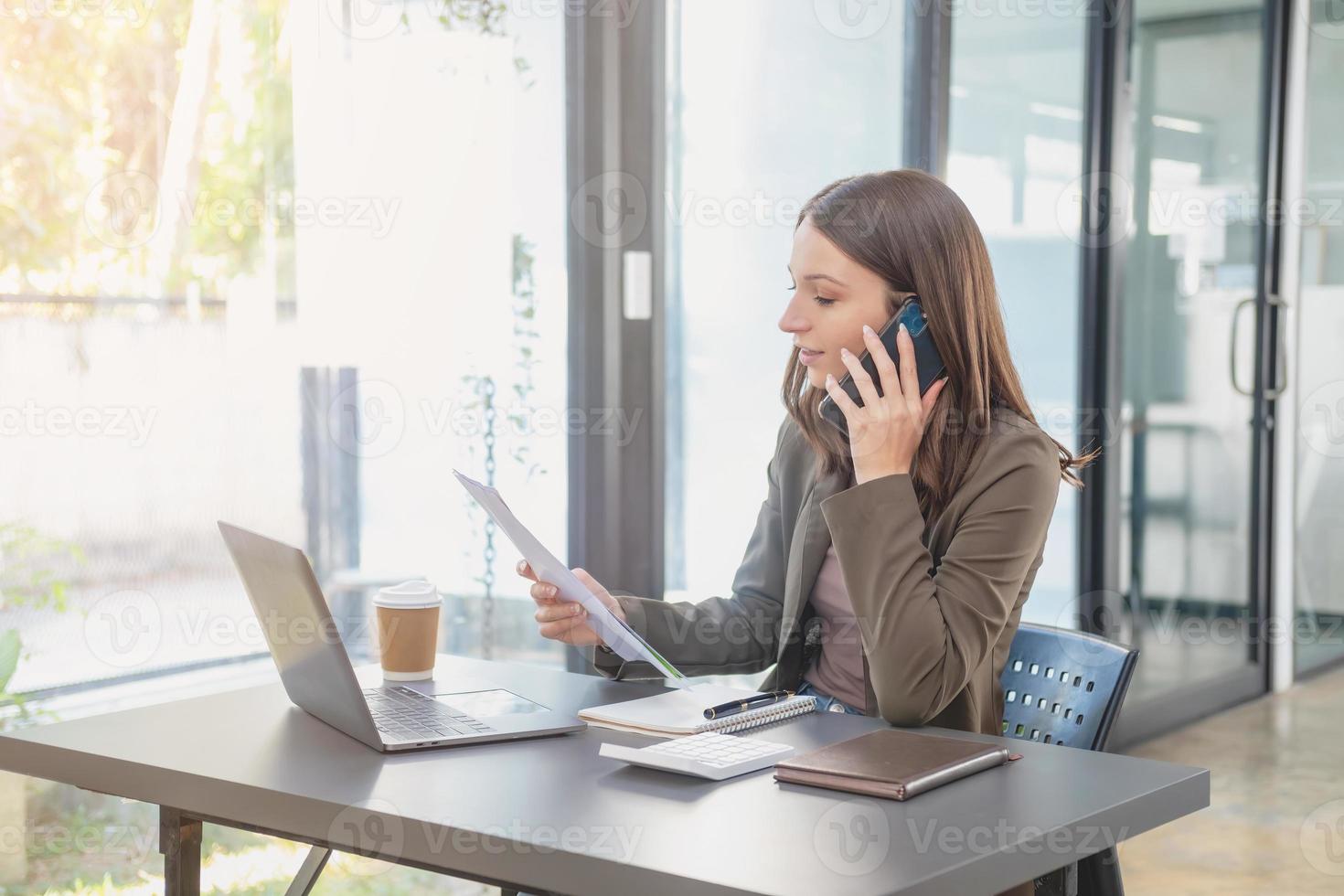 marketing, finanza, contabilità, pianificazione, donna d'affari di bi nazionalità è parlando con un' cliente che rappresentano un' azienda distributore utilizzando un' smartphone con il computer portatile penna e bloc notes su ufficio scrivania. foto