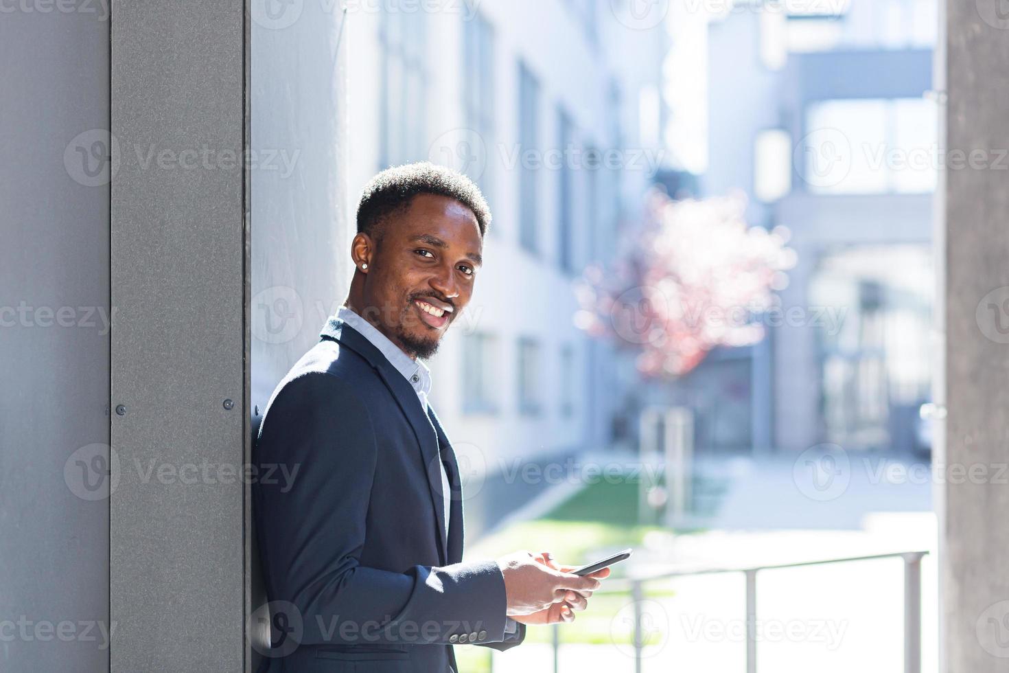 uomo d'affari afroamericano di successo e felice vicino all'ufficio che tiene il telefono cellulare foto