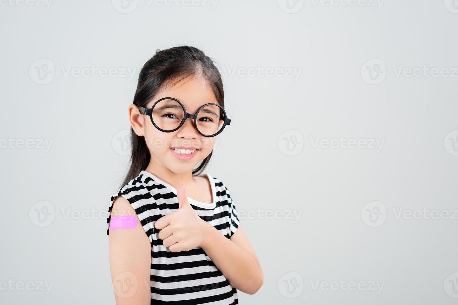 asiatico poco ragazza indossare bicchieri mostrando sua braccio con bendare dopo virus vaccino contento ragazzo e bambino vaccinazione campagna. proteggere il tuo ragazzo con vaccino concetto foto