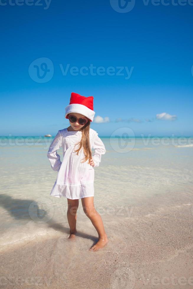 poco carino ragazza nel rosso cappello Santa Claus e occhiali da sole su il spiaggia foto