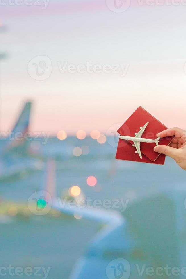 avvicinamento mano Tenere un aereo modello giocattolo e passaporti a il aeroporto sfondo grande finestra foto