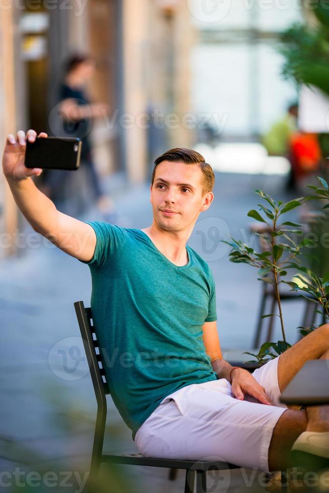 caucasico turista con smartphone assunzione autoscatto seduta nel all'aperto bar. giovane urbano ragazzo su vacanza esplorando europeo città foto