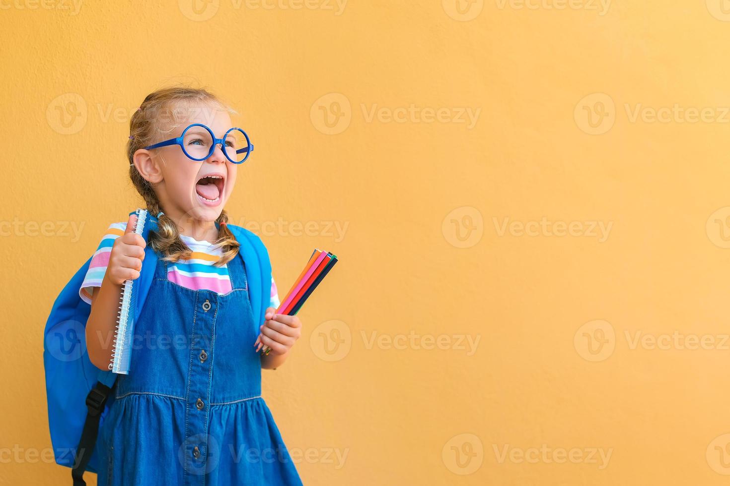 emotivo eccitato sorpresa viso scolara bambino nel bicchieri nel scuola uniforme detiene colorato matite, taccuino, zaino Aperto bocca per vuoto copia spazio attento isolato su giallo sfondo foto