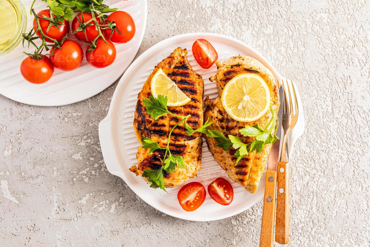 Due succoso grigliato pollo seni su un' bianca ceramica vassoio per servendo con verdure e Limone. superiore Visualizza. cemento grigio sfondo. delizioso cibo. foto