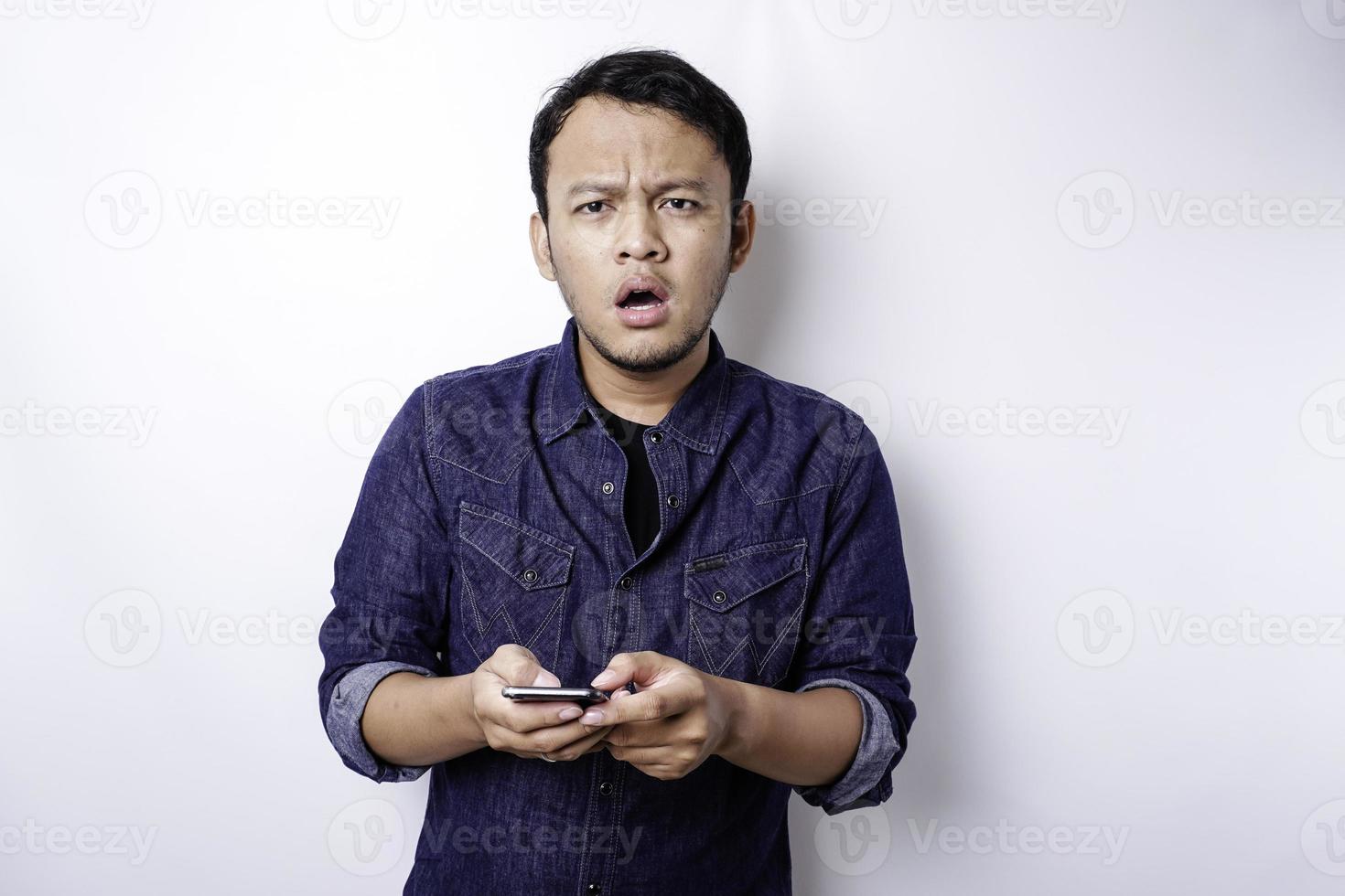 un' riflessivo giovane asiatico uomo è indossare blu camicia Tenere il suo Telefono e sembra confuso, isolato di bianca sfondo foto