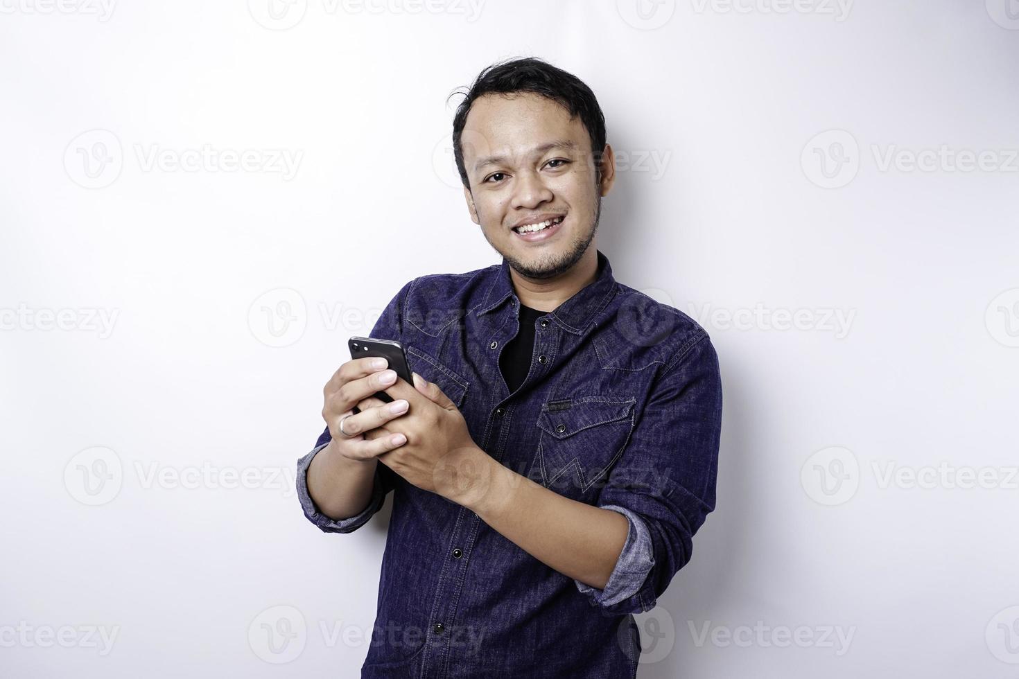 eccitato asiatico uomo indossare blu camicia sorridente mentre Tenere il suo Telefono, isolato di bianca sfondo foto