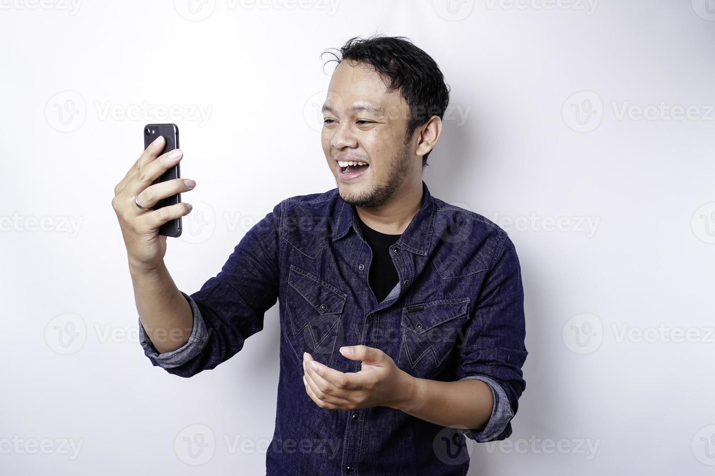 eccitato asiatico uomo indossare blu camicia puntamento a il copia spazio accanto lui mentre Tenere il suo Telefono, isolato di bianca sfondo foto