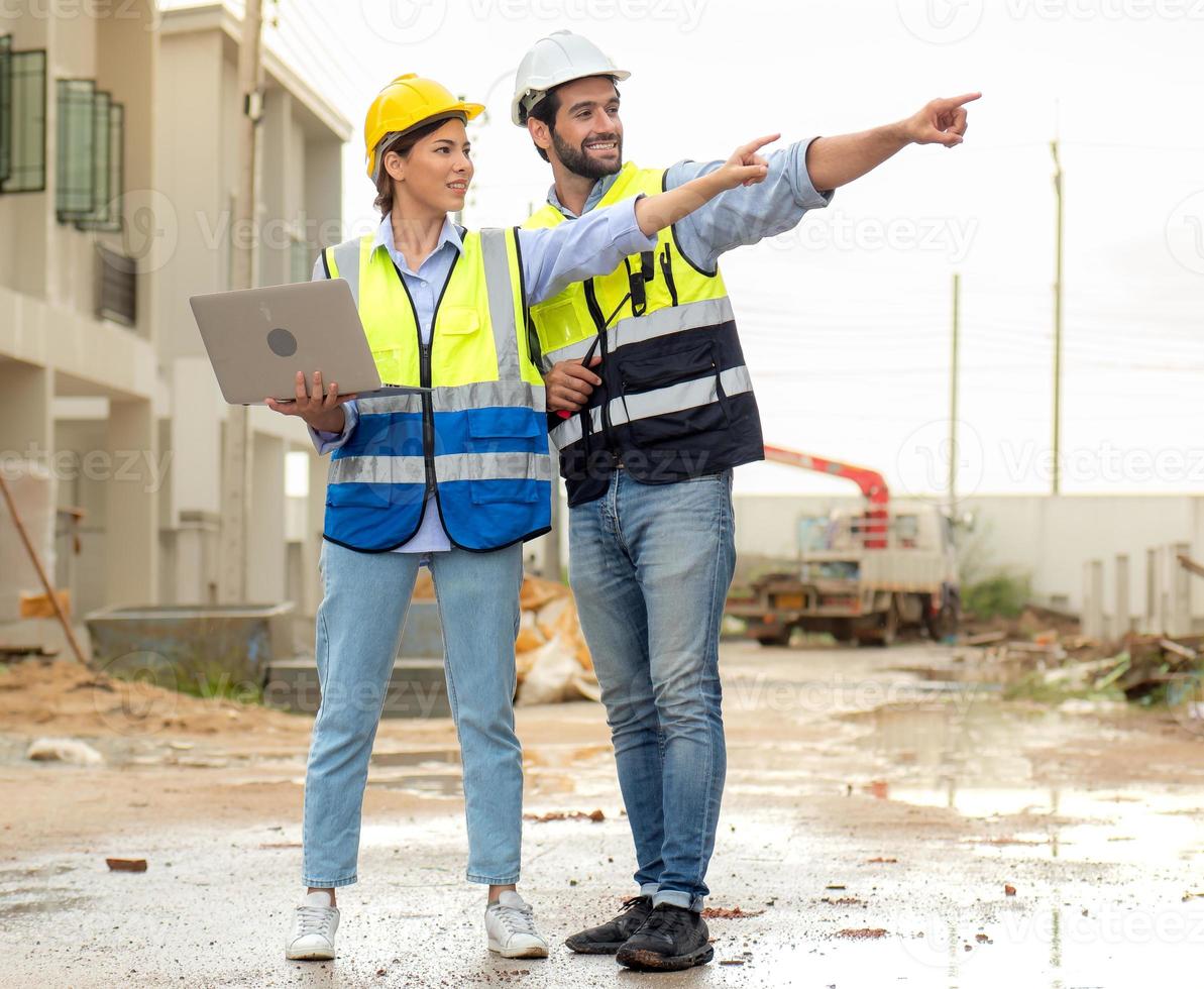 ingegnere uomo e femmina architetto indossare sicurezza caschi discutere alloggiamento sviluppo progetto a costruzione luogo utilizzando il computer portatile computer. contraente manager l'esame edificio tenuta infrastruttura. foto