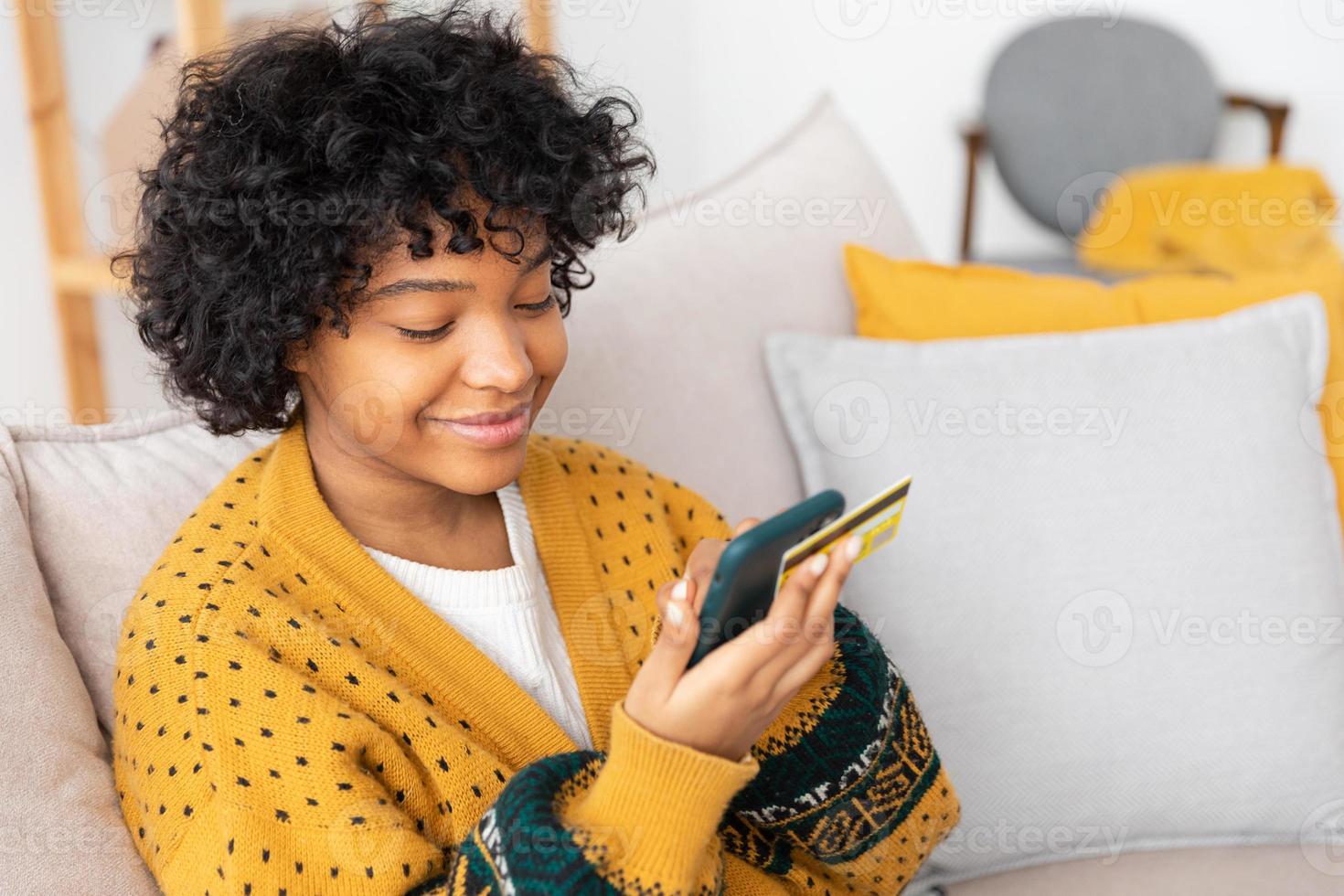 africano americano donna shopping in linea Tenere smartphone pagare con oro credito carta. ragazza seduta a casa acquisto su Internet accedere credito carta particolari. in linea shopping e-commerce consegna servizio. foto