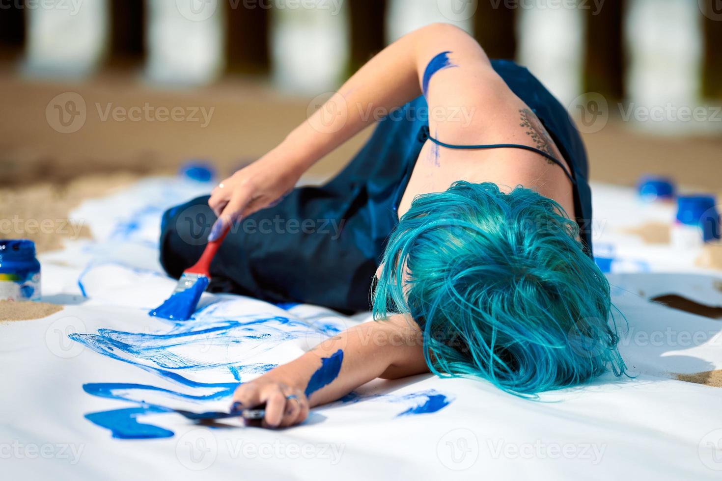 artista di performance artistica donna dai capelli blu imbrattata con pitture a guazzo su grande tela sulla spiaggia foto