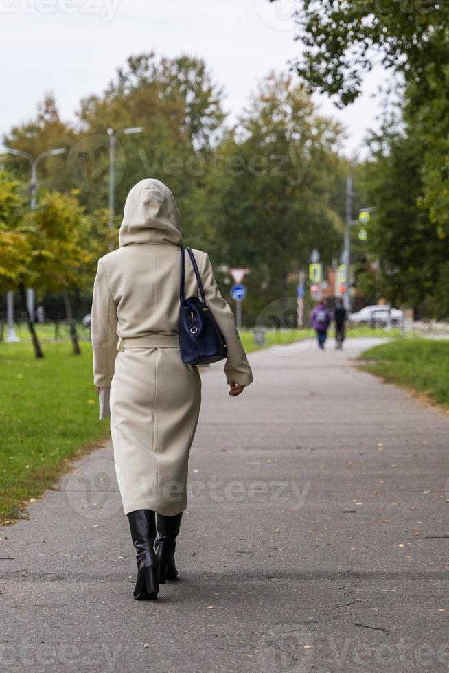 un' ragazza nel un' alla moda beige autunno cappotto, stivali con un' alla moda Borsa passeggiate lungo un' sentiero nel un' città parco foto